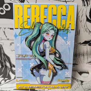 May include: A metal enamel pin featuring a character with long mint green hair, wearing a black and orange outfit, displayed on a rectangular backing card with the text REBECCA, Edgerunners, NIGHT CITY, and CYBERPUNK printed in yellow and white.