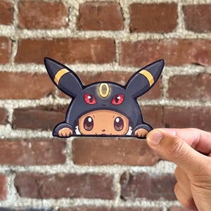 May include: A die-cut sticker featuring a tan creature wearing a dark purple Umbreon hood with yellow ring markings on the ears, red eyes, and a yellow forehead ring. The creature has large brown eyes and small paws visible at the bottom edge.