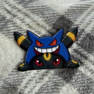 May include: A die-cut sticker featuring a black Umbreon character wearing a blue Gengar-themed hoodie. The design shows the Umbreon with red eyes and yellow rings, while the Gengar hood displays a wide, toothy grin and sharp, pointed ears.