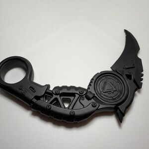 May include: A matte black karambit-style knife featuring a circular finger ring at the base, a textured handle with geometric cutouts, and a curved blade. The central circular section displays a triangular emblem with intricate mechanical detailing.