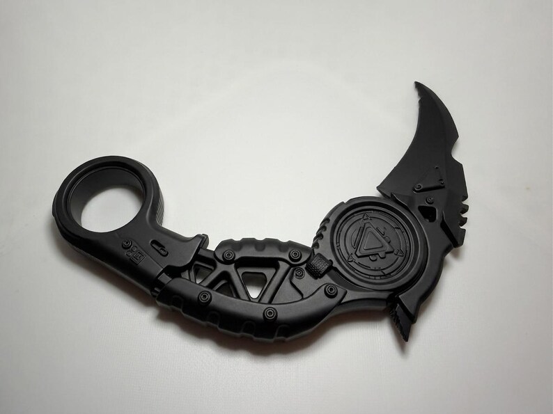 May include: A matte black karambit-style knife featuring a circular finger ring at the base, a textured handle with geometric cutouts, and a curved blade. The central circular section displays a triangular emblem with intricate mechanical detailing.