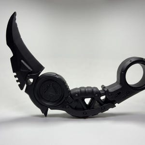 May include: A matte black karambit knife featuring a curved blade, a circular finger ring at the handle base, and a central circular emblem with a triangle design. The handle has a textured grip and mechanical detailing throughout the frame.