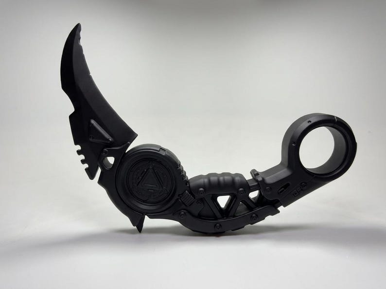 May include: A matte black karambit knife featuring a curved blade, a circular finger ring at the handle base, and a central circular emblem with a triangle design. The handle has a textured grip and mechanical detailing throughout the frame.