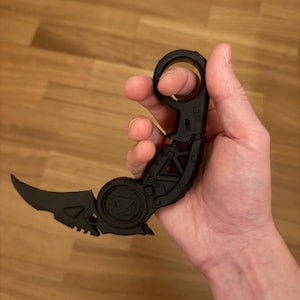May include: A matte black karambit-style knife featuring a curved blade, a textured handle with a circular finger ring at the pommel, and a mechanical-looking pivot point. The design includes geometric cutouts and a triangular emblem on the handle.