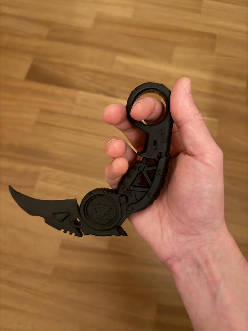 May include: A matte black karambit-style knife featuring a curved blade, a textured handle with a circular finger ring at the pommel, and a mechanical-looking pivot point. The design includes geometric cutouts and a triangular emblem on the handle.