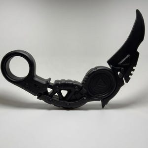 May include: A matte black karambit-style knife featuring a circular finger ring at the base, a textured grip section, and a curved blade. The handle includes a central circular emblem with a triangle detail, and the overall design is solid black.