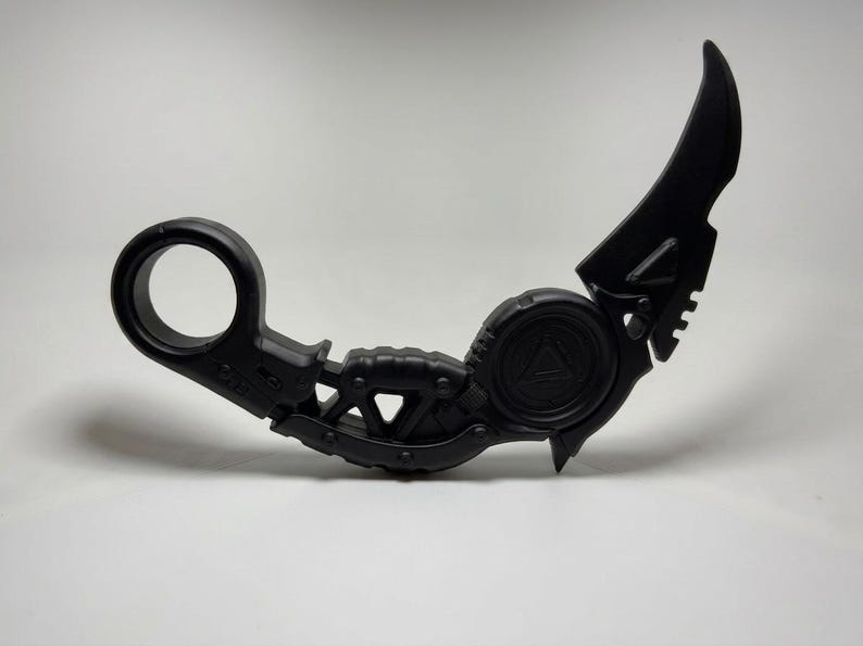 May include: A matte black karambit-style knife featuring a circular finger ring at the base, a textured grip section, and a curved blade. The handle includes a central circular emblem with a triangle detail, and the overall design is solid black.