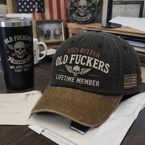 May include: A charcoal grey baseball cap with a brown brim featuring embroidered text that reads 100% BITTER OLD FUCKERS LIFETIME MEMBER with a skull and wings graphic. A small rectangular American flag patch is stitched onto the side panel.