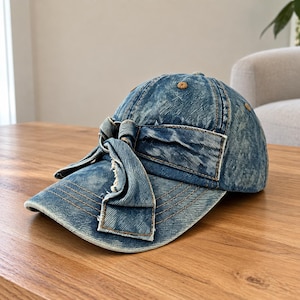 May include: A blue acid-wash denim baseball cap featuring a decorative fabric bow detail on the front panel. The cap has a curved brim with visible brown stitching and a metal ring accent integrated into the centre of the knotted denim bow.