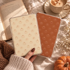 Puede incluir: Dos fundas rectangulares para teléfono con un patrón repetitivo de calabazas en líneas. Una funda tiene fondo crema con contornos marrones, la otra tiene fondo naranja quemado con contornos blancos. Ambas fundas son transparentes.