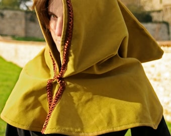 May include: A mustard-colored medieval hood with a pointed tip and a brown braided trim. The hood is tied at the neck with a matching brown cord. The person is wearing a black top.