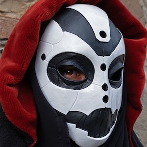 May include: A person wearing a black hooded cloak with a red lining. The person is wearing a white and black mask with a geometric design.