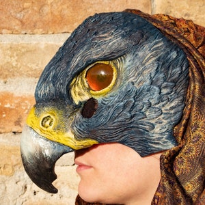Könnte beinhalten: Eine Person trägt einen braunen und goldenen gemusterten Schal mit einer blauen und gelben Vogelmaske, die ihr Gesicht bedeckt. Die Maske hat ein realistisches Design mit einem großen Schnabel und einem einzigen Auge.