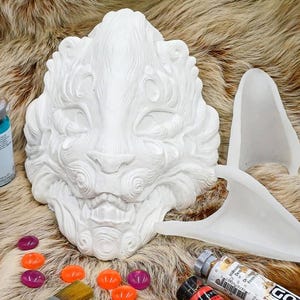 Puede incluir: Máscara de cerámica blanca con detalles intrincados, que se asemeja a una criatura mítica, con pinceles, tubos de pintura y cuentas de vidrio de colores sobre una superficie de piel sintética.