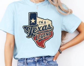 May include: A light blue short-sleeve t-shirt featuring a distressed graphic of the state of Texas in red, white, and blue. The graphic includes a white star and the text I'M A Texas LEGEND in a vintage-style font across the centre of the state.