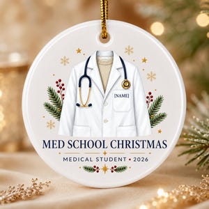 May include: A round white ceramic ornament featuring a graphic of a white lab coat over a beige sweater with a gold and blue stethoscope. The text reads MED SCHOOL CHRISTMAS, MEDICAL STUDENT, 2026, and [NAME]. Decorated with pine and berries.