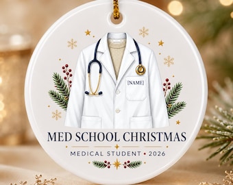 May include: A round white ceramic ornament featuring a graphic of a white lab coat over a beige sweater with a gold and blue stethoscope. The text reads MED SCHOOL CHRISTMAS, MEDICAL STUDENT, 2026, and [NAME]. Decorated with pine and berries.