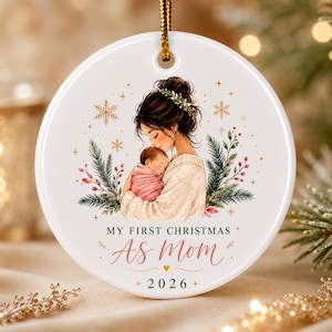 May include: A round white ceramic Christmas ornament featuring an illustration of a person holding a baby wrapped in a pink blanket. The text reads MY FIRST CHRISTMAS AS MOM 2026, surrounded by pine branches, red berries, and gold snowflakes.