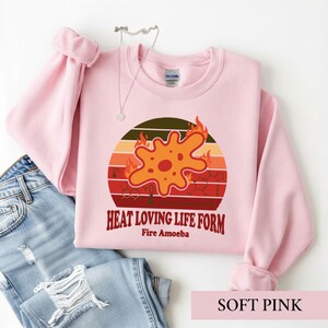 May include: A light pink crewneck sweatshirt featuring a graphic of an orange amoeba surrounded by flames against a sunset-style background with chemical structures and the text "HEAT LOVING LIFE FORM" and "Fire Amoeba" printed in dark red.