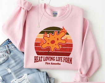 May include: A light pink crewneck sweatshirt featuring a graphic of an orange amoeba surrounded by flames against a sunset-style background with chemical structures and the text "HEAT LOVING LIFE FORM" and "Fire Amoeba" printed in dark red.