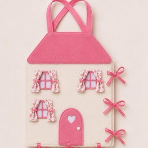 May include: A house-shaped felt wall hanging in cream and pink. It features a pink roof, three windows with floral curtains, and a pink door with a heart cutout. The side and bottom edges are decorated with pink ribbon bows and hanging loops.