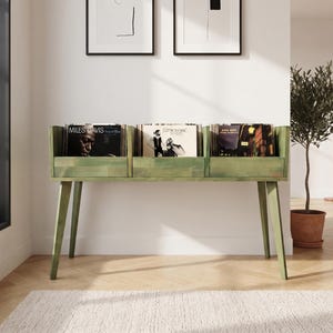 Può includere: Una console in legno verde chiaro per riporre dischi in vinile, dotata di tre scomparti aperti e sostenuta da quattro gambe inclinate. L'unità contiene diversi album in vinile, inclusi titoli di Miles Davis, disposti in posizione verticale.