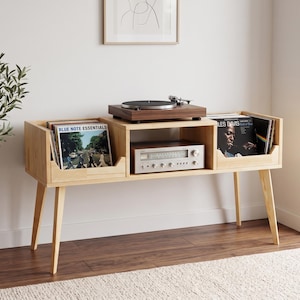 Op de afbeelding: Een lichtgekleurd houten mediameubel met vier taps toelopende poten, twee zijvakken voor vinylplaten en een centrale plank met een zilverkleurige audio-ontvanger. Een donkerhouten platenspeler staat op het bovenoppervlak van het meubel.