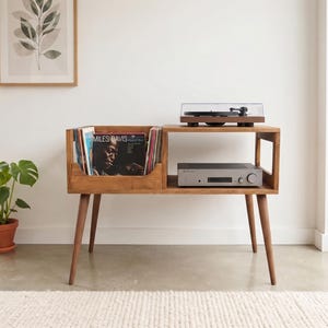 May include: A wooden record player console with four tapered legs, featuring a left-side storage compartment for vinyl records and a right-side shelf holding a silver audio amplifier, topped with a black turntable under a clear cover.
