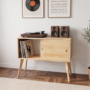 May include: A light-colored wooden media console with four tapered legs, featuring an open storage shelf on the left side containing vinyl records and a sliding door cabinet on the right side, topped with a dark wood record player and two speakers.