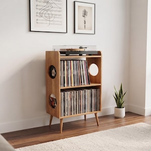 Puede incluir: Un mueble de madera de color marrón claro para almacenar discos de vinilo con cuatro patas cónicas. La unidad cuenta con dos estantes abiertos llenos de discos, una superficie superior con un tocadiscos y aberturas circulares laterales.