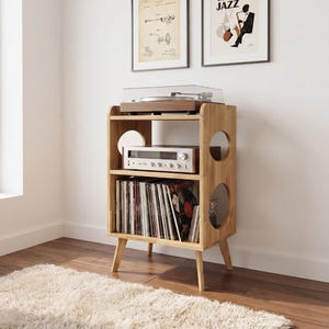Peut inclure: Un meuble multimédia en bois clair à trois niveaux de rangement. L'étagère supérieure accueille une platine vinyle, l'étagère centrale contient un amplificateur argenté et l'étagère inférieure est remplie de disques vinyles. Il a des découpes circulaires.