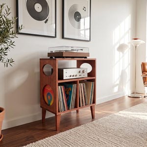 Puede incluir: Un mueble de madera de color marrón rojizo para tocadiscos, con un estante superior para el reproductor, un estante central para el amplificador y dos compartimentos inferiores para discos de vinilo, con aberturas circulares en los laterales.