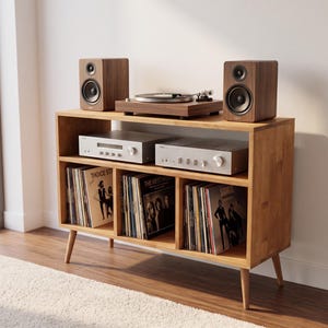 May include: A light brown wooden media console with four tapered legs, featuring an open top shelf holding a turntable and two speakers, a middle shelf with two silver audio components, and three lower compartments filled with vinyl records.