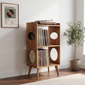May include: A brown wooden record storage cabinet with two open shelves filled with vinyl records. The unit features four tapered legs and circular side cutouts. A turntable sits on the top surface of the cabinet, which is positioned in a room.
