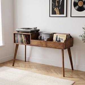 Puede incluir: Una mesa consola de madera marrón con cuatro patas cónicas, que presenta una superficie superior plana con un tocadiscos y dos compartimentos de almacenamiento abiertos llenos de discos de vinilo dispuestos verticalmente en el marco.