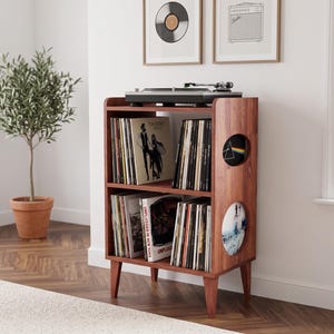 May include: A brown wooden record player stand with two shelves filled with vinyl records. The unit features four tapered legs and two circular side cutouts. A black turntable sits on the top surface, and the shelves are organized with various album covers.