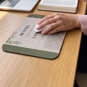 May include: A rectangular mouse pad featuring a light wood grain background, green leaf accents, and the printed text BE STILL AND KNOW THAT I AM GOD. The mouse pad is positioned on a wooden desk next to an open book, a laptop, and a white mug.