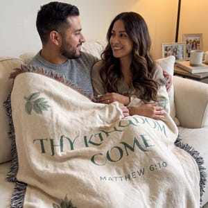 May include: A cream-coloured woven throw blanket featuring green leaf illustrations and the text "THY KINGDOM COME" followed by "MATTHEW 6:10" in dark green lettering. The blanket has a decorative fringe edge with multi-coloured threads.