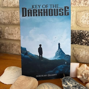 May include: A paperback book titled "Key of the Darkhouse" by Deborah Zelazny. The cover features a blue sky with clouds, a silhouette of a person standing on a rocky cliff, and a lighthouse in the distance.