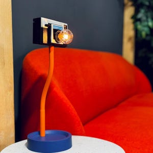 Op de afbeelding: Een unieke bureaulamp in de vorm van een vintage camera. De lamp heeft een zwart en zilveren camerabody met een gloeilamp als lens. Hij heeft een oranje steel en een blauwe voet, geplaatst op een witte tafel. De achtergrond is een rode bank.