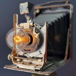 Puede incluir: Una cámara vintage con una bombilla conectada en la parte delantera, montada en un trípode. La cámara tiene un sistema de fuelle y un visor en la parte superior. La bombilla está encendida.