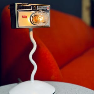 Könnte beinhalten: Eine Vintage Kodak Instamatic 104 Kamera, die als Lampe umfunktioniert wurde. Die Kamera ist auf einem weißen, wellenförmigen Metallständer mit rundem Sockel montiert. Die Glühbirne ist im Kameraobjektiv sichtbar. Die Lampe steht auf einem grauen Tisch.