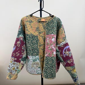 May include: A long-sleeve quilted jacket featuring a patchwork design with various floral patterns in shades of mustard yellow, forest green, burgundy, and cream. The jacket has a round neckline, an open front, and a visible orange brand label.