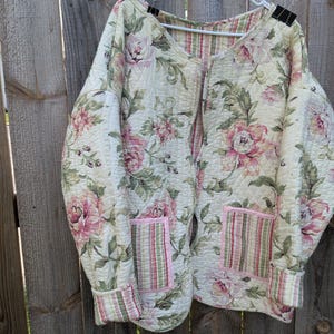 May include: A quilted jacket featuring a cream base with a floral pattern of pink roses and green leaves. The garment includes two front pockets with a pink, green, and cream striped design, matching the cuffs and the interior lining visible at the neck.