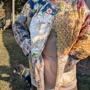 May include: A long-sleeve quilted jacket featuring a patchwork design with various floral and botanical patterns in shades of yellow, blue, green, and red. The jacket has a collarless neckline, a front opening, and two visible side pockets.