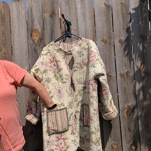May include: A long-sleeved quilted jacket featuring a cream base with a pink and green floral pattern. The jacket has a round neckline, an open front, and two large patch pockets on the front that display a contrasting vertical striped fabric.