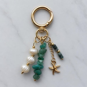 May include: A gold-toned metal keyring featuring a circular clasp with three dangling charms: a strand of three white pearls, a strand of raw green stone chips, and a gold-toned starfish charm paired with small dark green and gold beads.