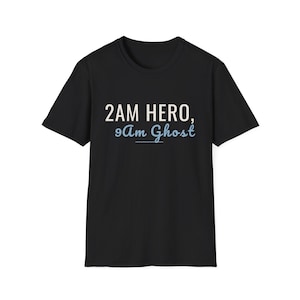 May include: A black short-sleeved t-shirt featuring the text 2AM HERO, in white capital letters, followed by the text 9Am Ghost, in light blue script font with an underline, centered on the chest area of the garment.