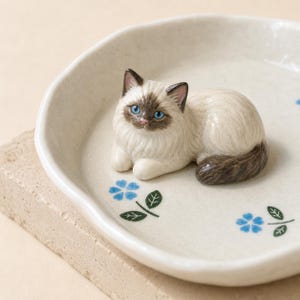 May include: A ceramic trinket dish featuring a sculpted, cream-colored cat with dark brown points and bright blue eyes resting in the center. The dish has a speckled off-white finish and is decorated with small, painted blue flowers and green leaves.