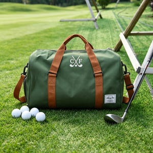 May include: An olive green duffel bag with brown straps and a white embroidered logo featuring crossed golf clubs and the letters C and F. The bag has a small rectangular white label in the bottom right corner and is surrounded by four white golf balls.